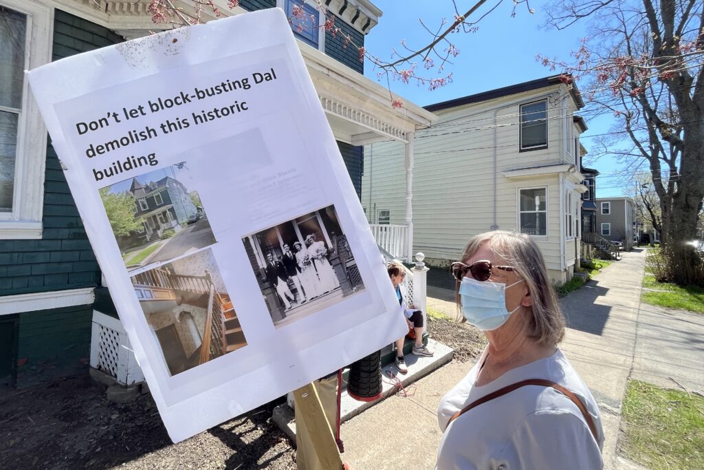 一名戴着口罩的女人和太阳镜在住宅城市人行道上拿着标志。该标志包括那个女人背后的房屋照片，上面写着：“不要让破坏性的达尔拆除这座历史悠久的建筑”。