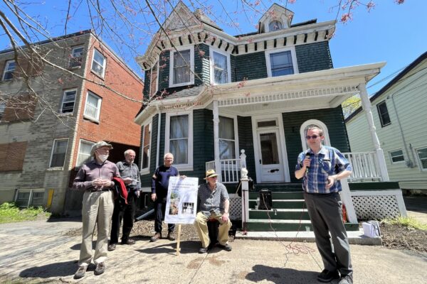 A man wearing glasses and a short-sleeve plaid shirt speaks into a microphone on a sunny day. Behind him is a group of men, one holding a sign, and an old house. The house is forest green with white trim.