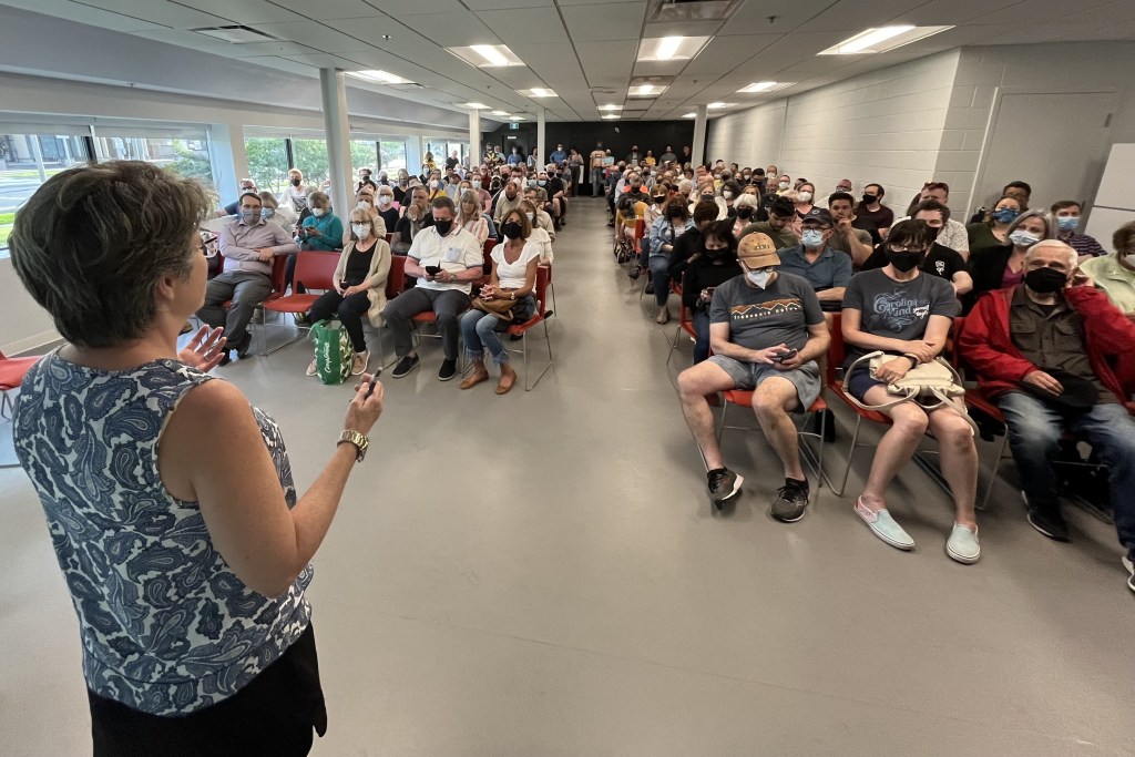 一个穿着蓝白相间的佩斯利衬衫的女人对着一屋子坐在红色椅子上的人说话，他们中的许多人都戴着面具。