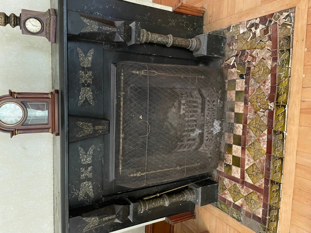 A black fireplace with ornate details is shown inside an old house. There's a grate in front of the fireplace, and multi-coloured tile on the floor in front. The tile is smashed. On the mantle there are two old clocks.