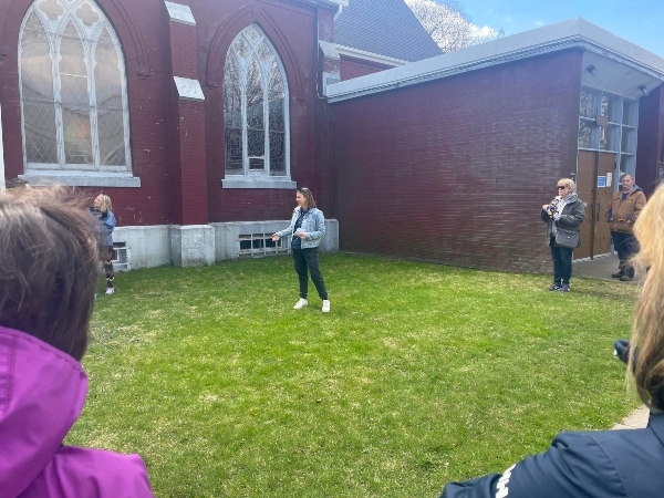 一名身穿浅色牛仔夹克、黑色裤子和白色运动鞋的女子站在红砖教堂附近的草地上与人们交谈。