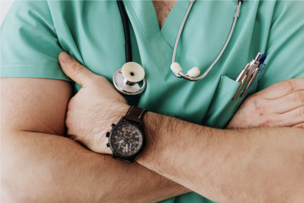 一名穿着绿色手术服的男医生的胸部照片，胸前放着听诊器，双手交叉放在胸前，展示他的手表。