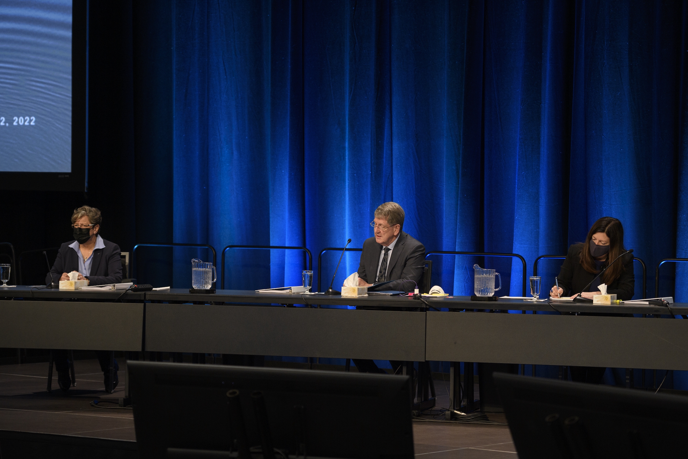 三个戴着口罩的人坐在一张桌子前，前面是蓝色的窗帘