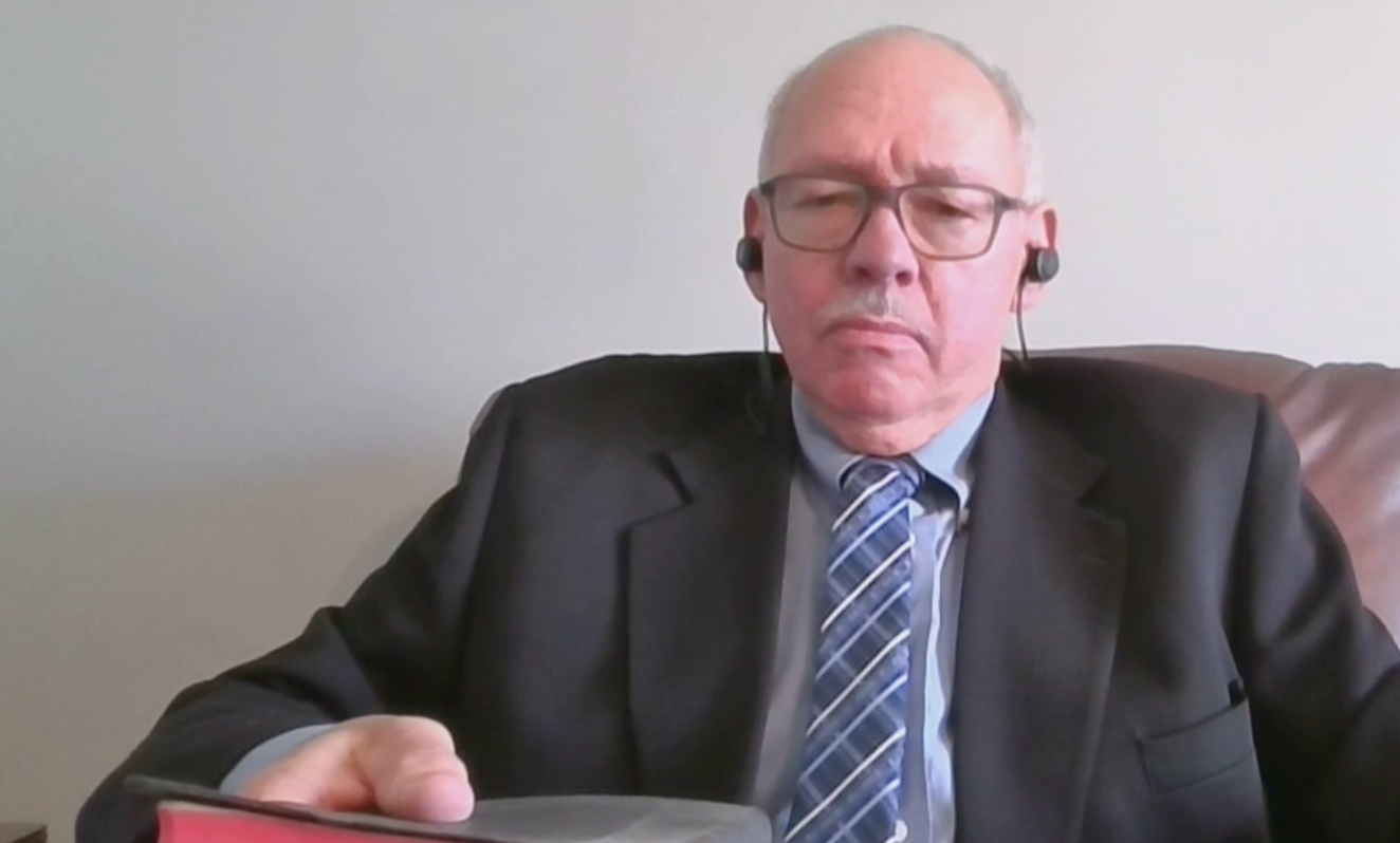 一个秃顶的男人戴着眼镜，穿着深色套装，打着蓝色图案领带，坐在电脑前参加Zoom会议