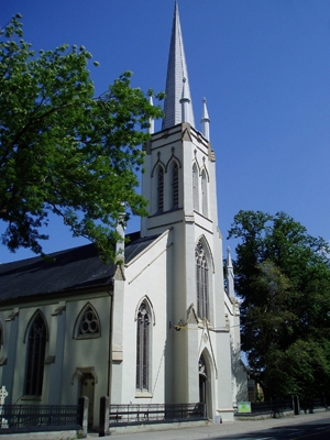 A pale grey stone church with a tall steeple is surrounded by a black iron fence, tall green trees, and a blue sky.