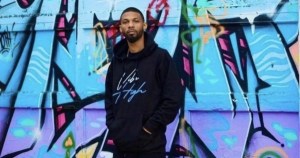 一个穿着黑色夹克和裤子的黑人站在一幅彩色的蓝色壁画前，壁画上有多种颜色的图案