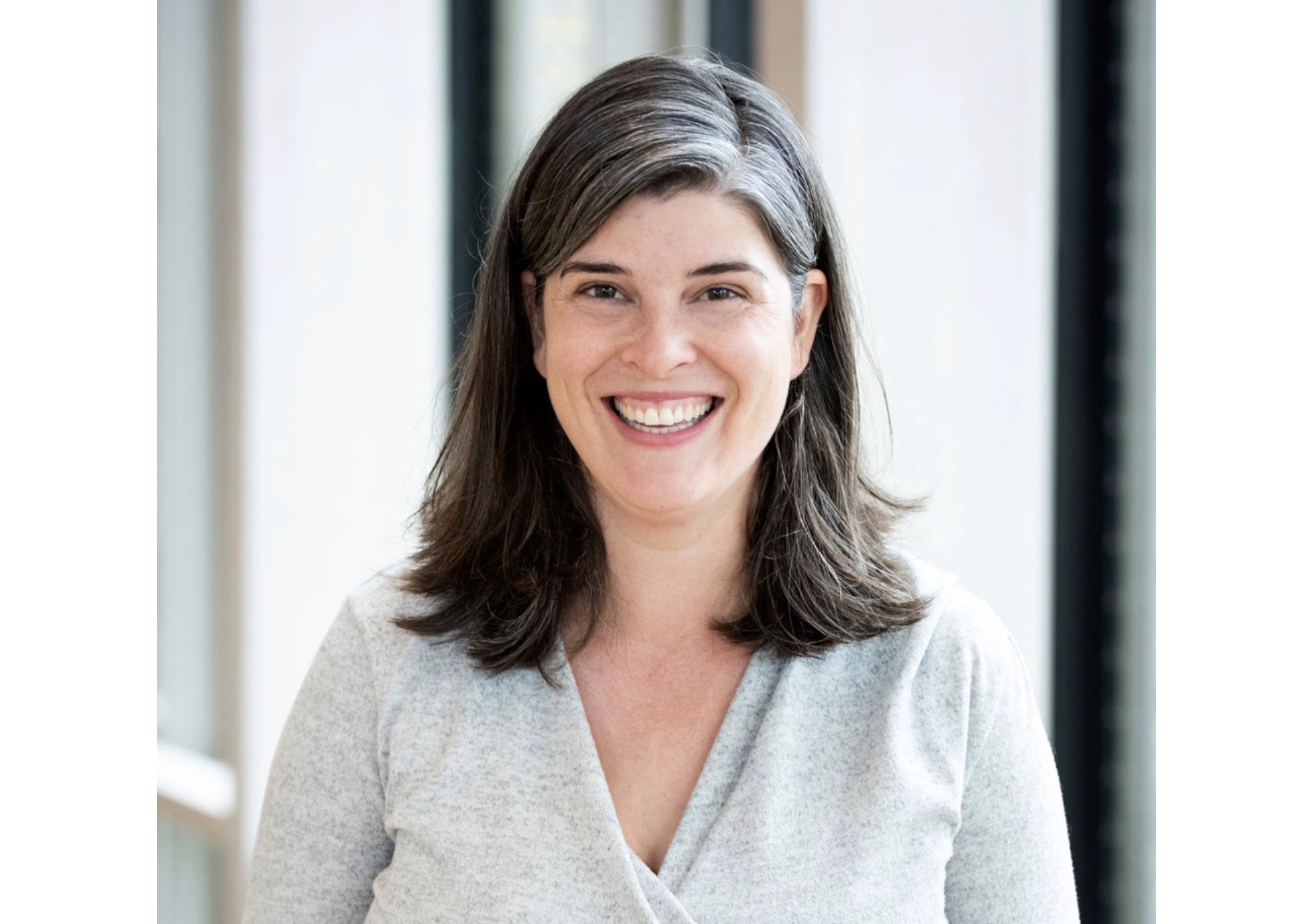 一个穿着浅灰色长袖衬衫的女人微笑着站在一排窗户旁。