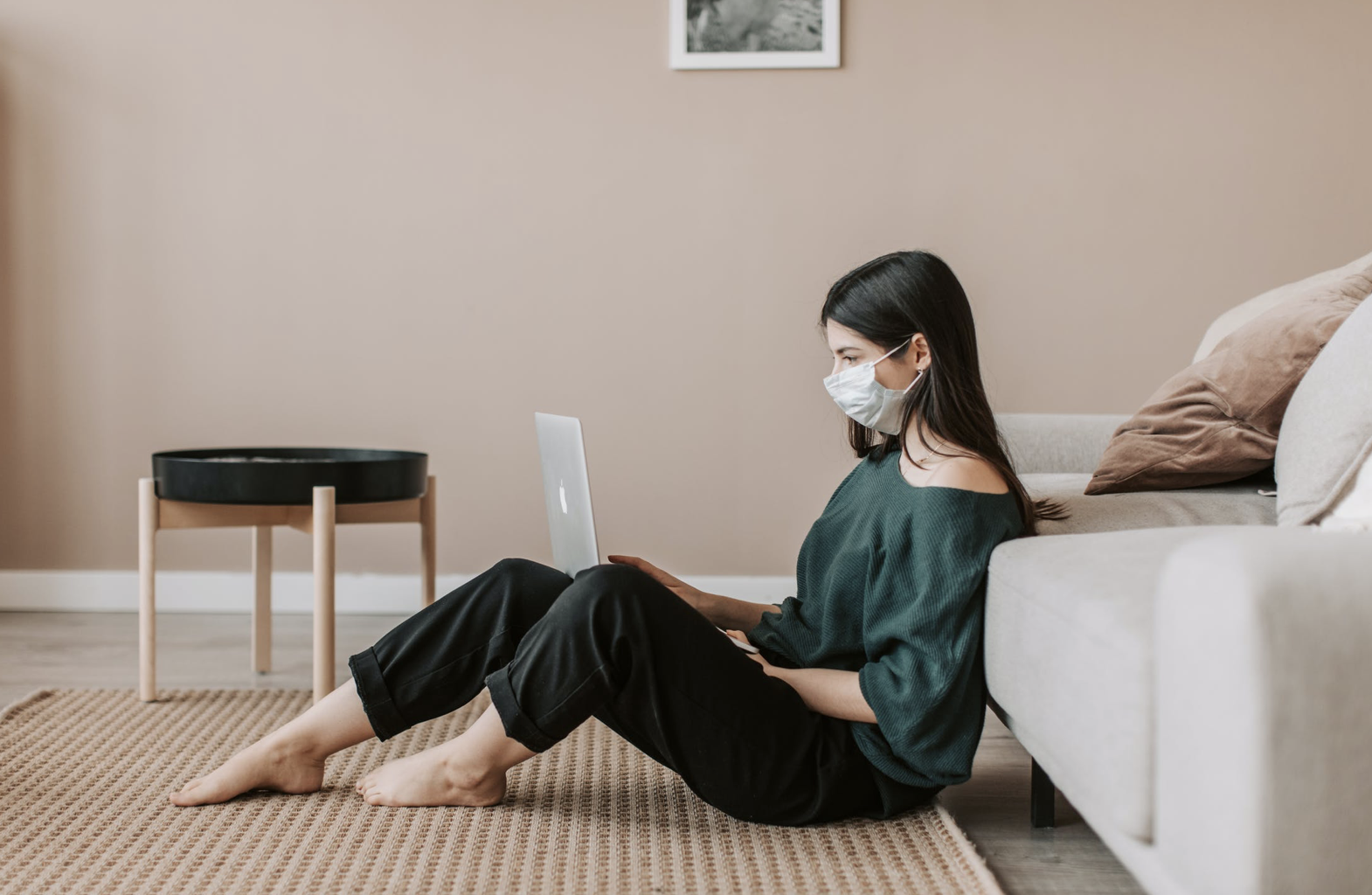 一个年轻女子坐在客厅的地板上，靠在米色沙发上，戴着口罩，同时在笔记本电脑上。