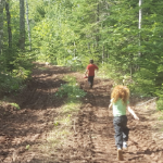 Two kids run on an access road in a bright green forested area