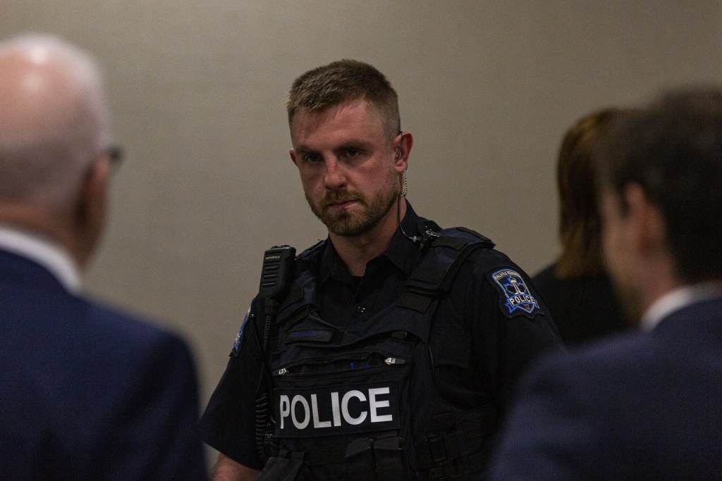 一名身穿警察制服的男子走进一间灯光昏暗的米黄色房间。他看着一个穿西装、头发灰白的男人，右边还有一个穿西装、头发乌黑的男人。