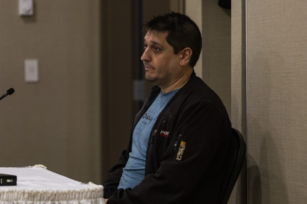 一个男人穿着一件蓝色的t恤和黑色夹克圣ares ahead. He's sitting in a beige room, with a microphone and bible on a table in front of him.