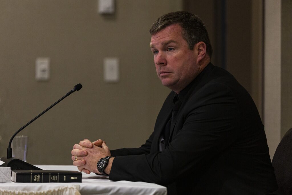 一个穿着全黑西装和手表的男人向前看，向左望着。他在一个米色的房间里，他面前的桌子上有一个麦克风和圣经。