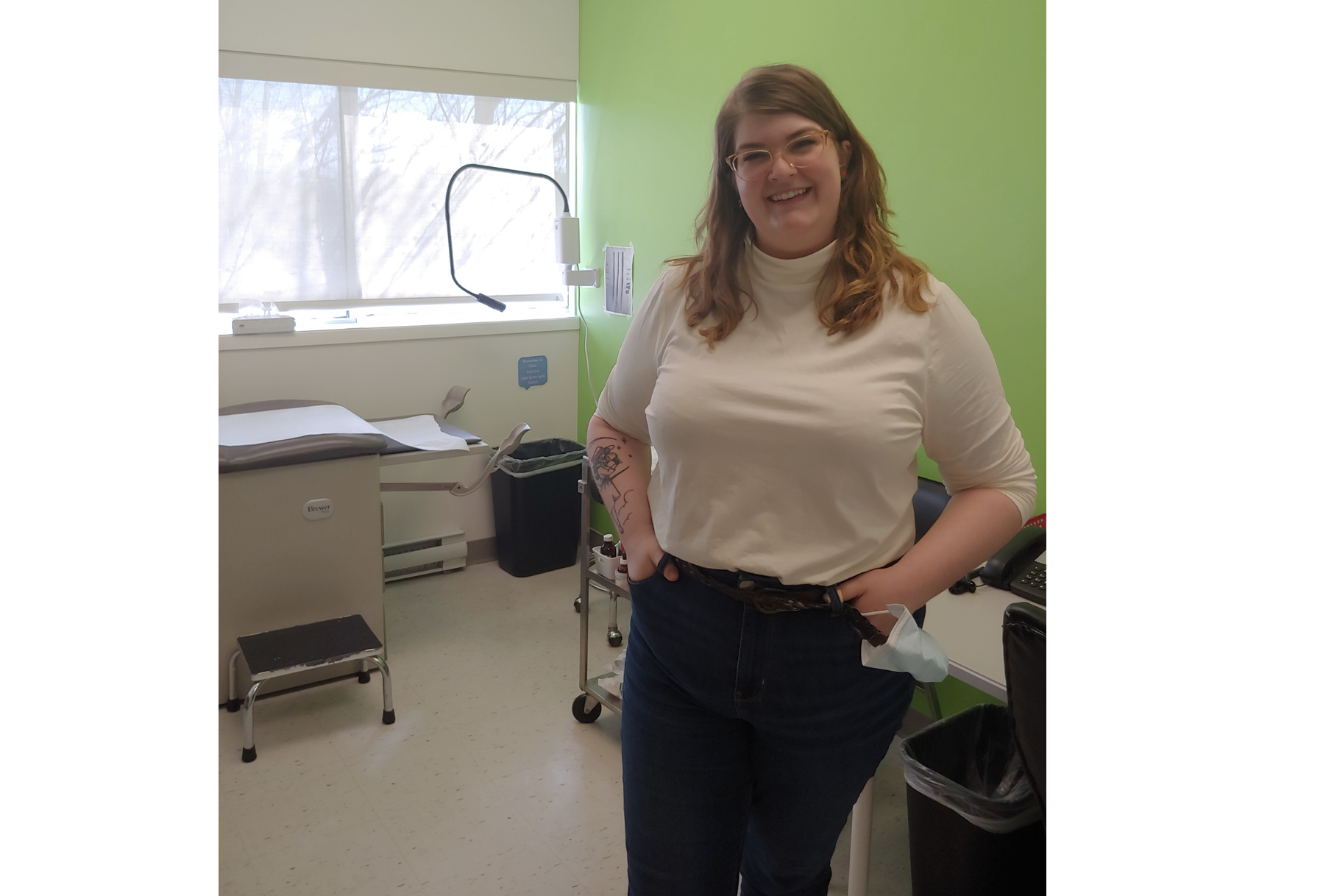 在绿色墙壁的医务室里，一位身穿长袖白衬衫和深色裤子、面带微笑的妇女靠在一张桌子上。