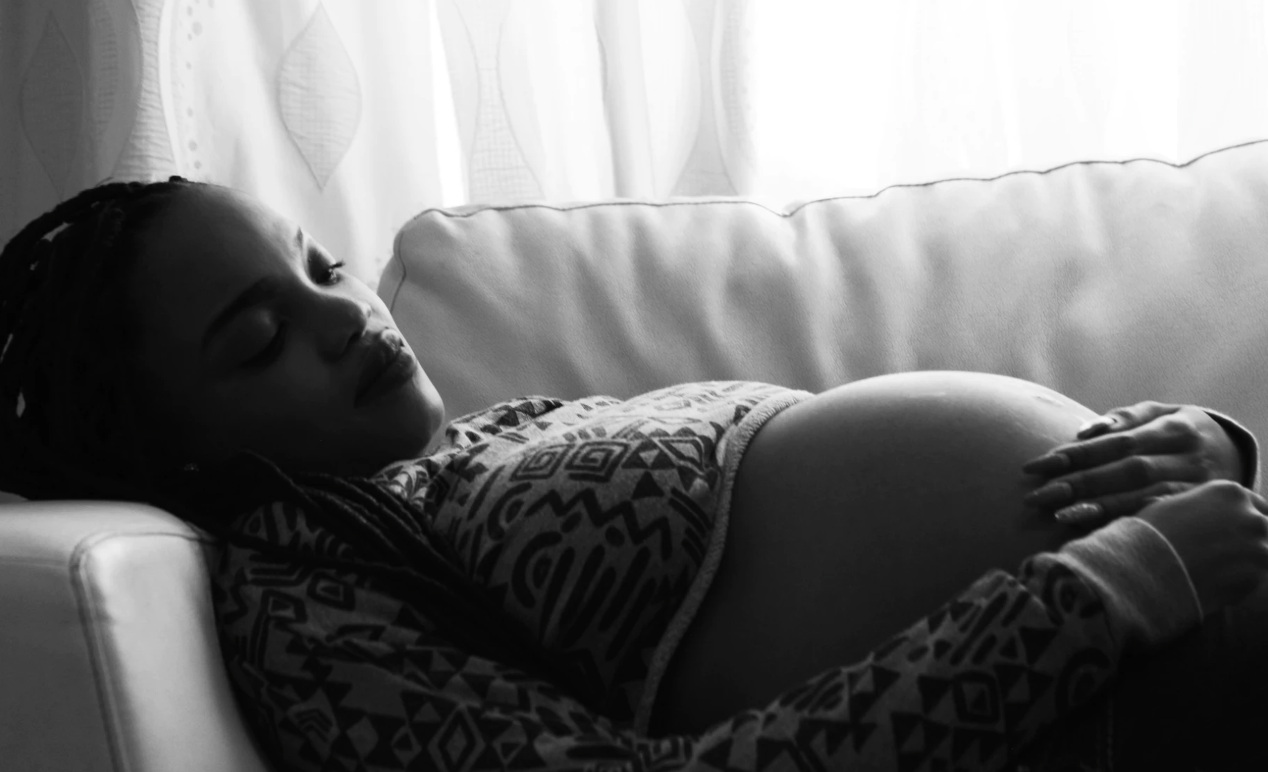 一张黑白照片上，一位孕妇躺在窗户前沙发的阴影里，双手捂着肿胀的腹部，低头沉思。