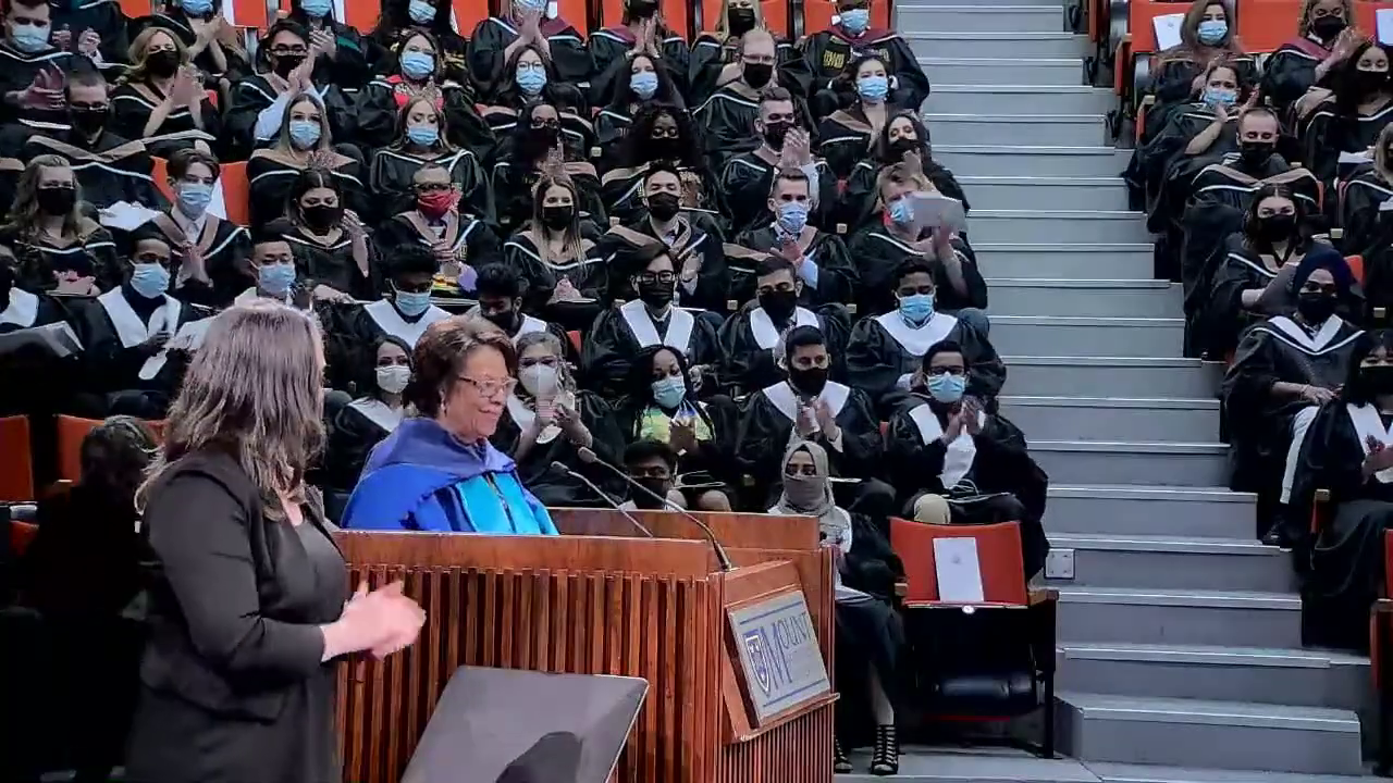 身穿蓝色毕业服的黑人女士在讲台上发表演讲，身穿黑色毕业服的学生在背后鼓掌