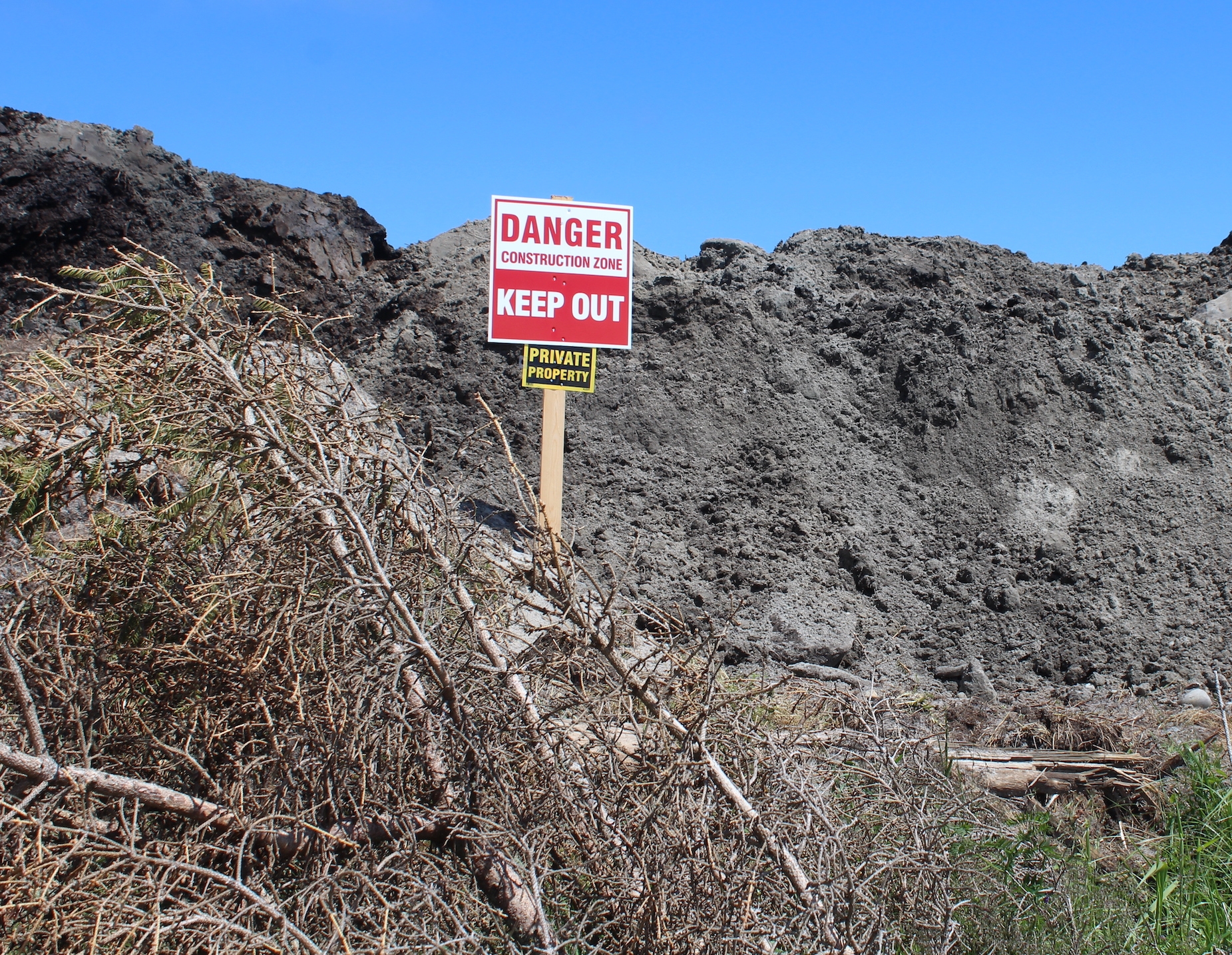 一个红白相间的牌子写着“危险，施工区域，请勿入内”，另一个黑黄相间的牌子写着“私人财产”。这些标志放在一堆泥土和折断的树枝上。