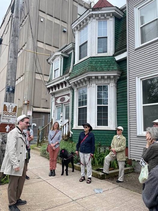 一小群人站在一座绿色的两层楼房子前的人行道上，房子有白色的镶边和凸窗。