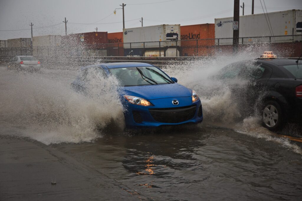 一辆蓝色的汽车在黑暗的日子里溅起了巨大的水花。从蓝色车辆流出的水浸透了一辆向相反方向行驶的出租车的挡风玻璃。在这些车辆的后面是一系列集装箱，它们位于与道路平行的火车轨道上。