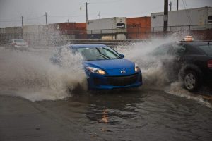 一辆蓝色的汽车在黑暗的日子里溅起了巨大的水花。从蓝色车辆流出的水浸透了一辆向相反方向行驶的出租车的挡风玻璃。在这些车辆的后面是一系列集装箱，它们位于与道路平行的火车轨道上。