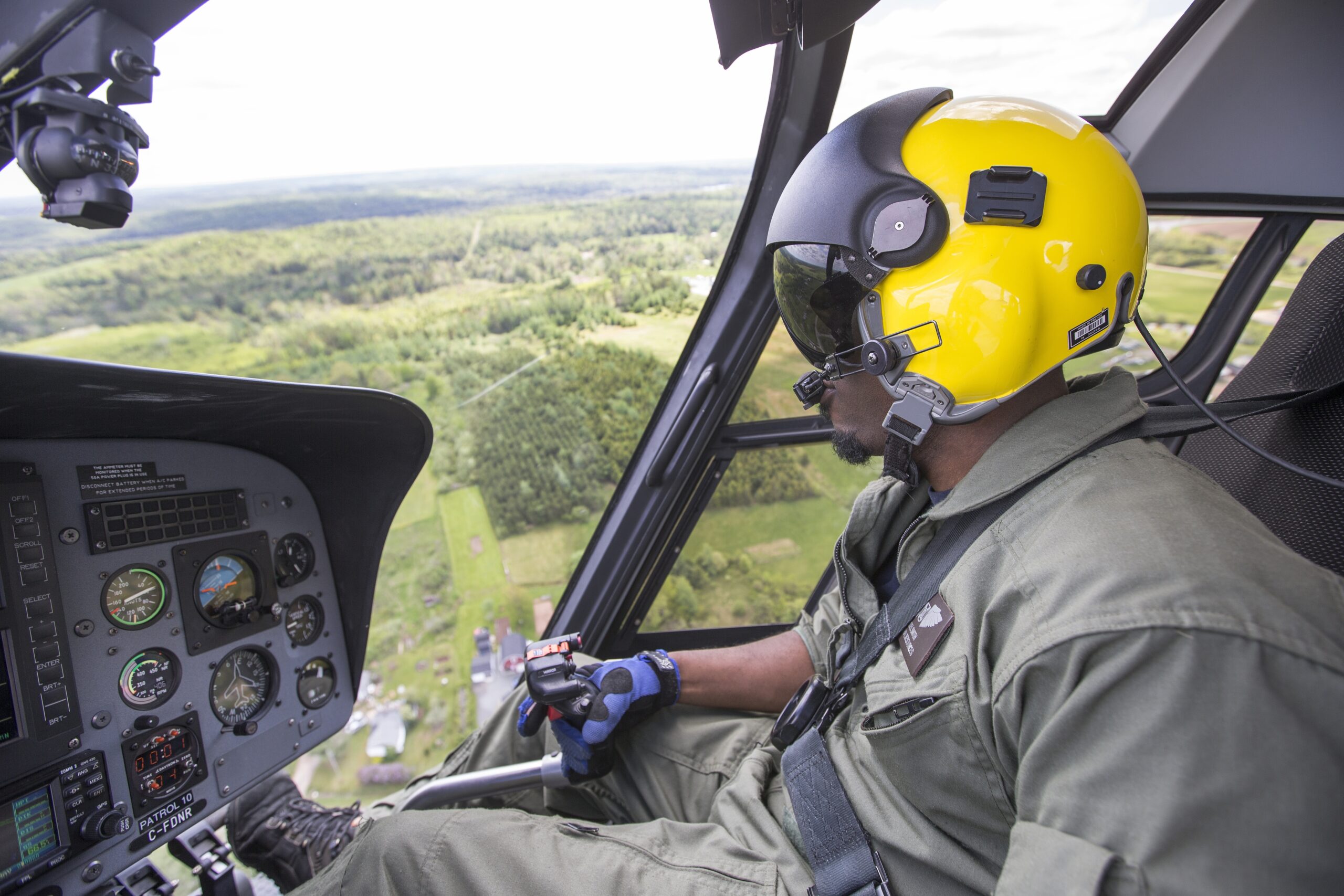 Side view of a Black man in a yellow helmet flying a helicopter with green land in the background