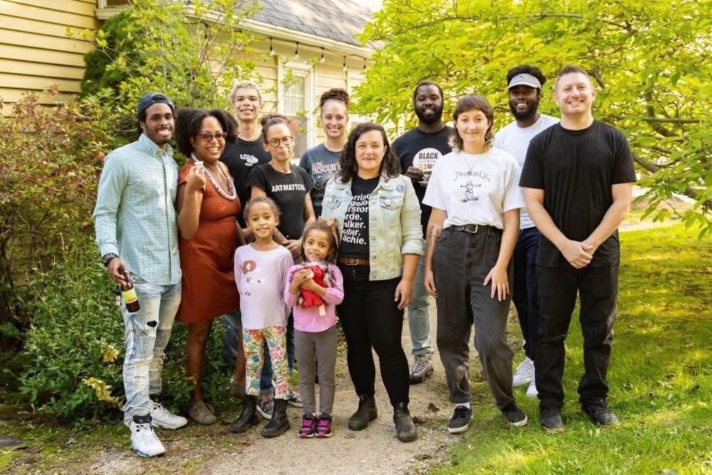 A group of people stand out in a yard in front of a yellow historic house surrounded by lush trees and greenery.