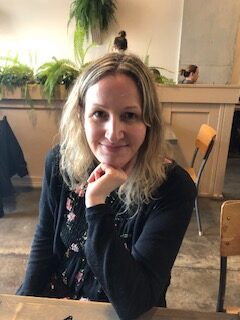 一位留着齐肩卷发、穿着黑色毛衣的女士坐在桌子旁，手放在下巴下。