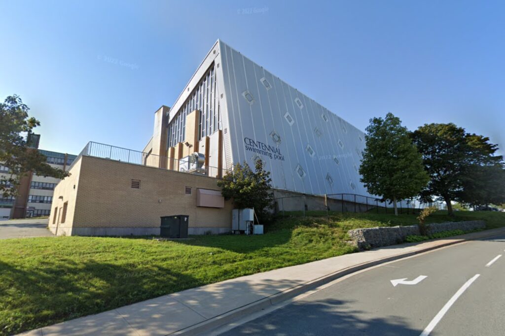 An arched, midcentury building with white cladding on one side and beige brick on the other, is seen from the road on a sunny day.