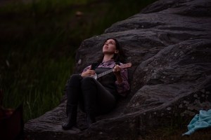 A woman with dark hair, a purple top, and black overalls is lying on a rock and playing the banjolele.