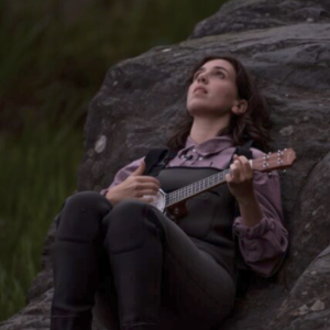 黄昏时分，一位黑发紫衫的年轻白人女子躺在一块大石头上，一边仰望天空，一边弹奏着她的班乔莱。