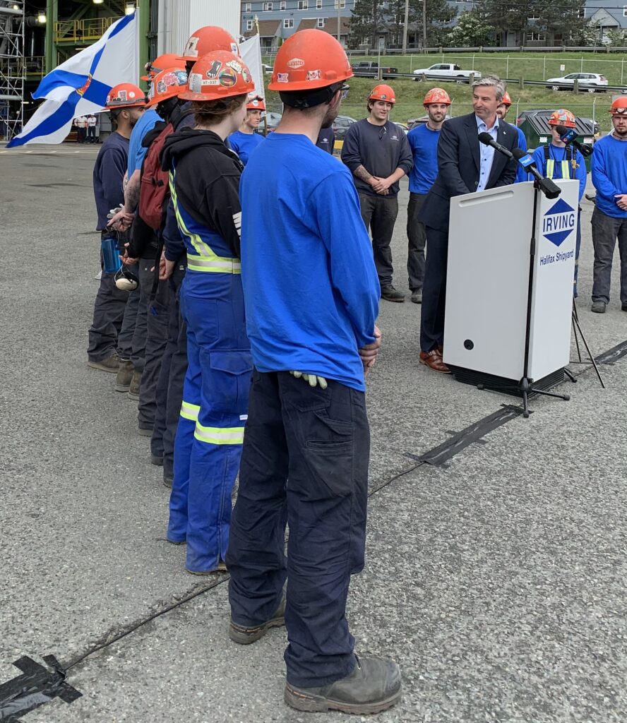 一个穿着盐和胡椒头发的男人戴着西装的架子在船厂的讲台上，被硬帽和工作服的商人包围。