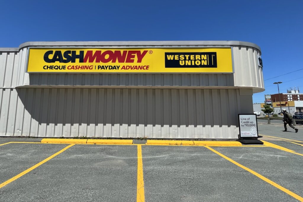 A grey building with a yellow sign reading 'CASHMONEY CHEQUE CASHING PAYDAY ADVANCE,' is seen on a sunny day.