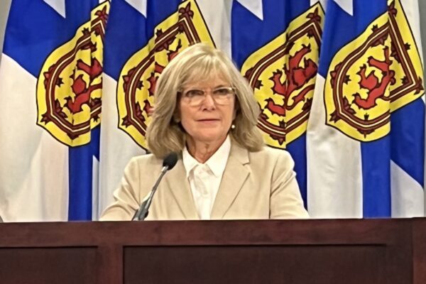 在一张模糊的手机照片中，一位女士坐在麦克风前。她穿着白色衬衫和米色运动夹克。在她身后是四面新斯科舍省国旗——白色带有蓝色对角线，黄色的顶部有一只红色的狮子。