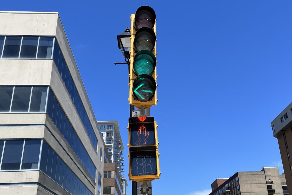 在阳光明媚的日子里，从低角度看红绿灯。交通方面，有一个普通的绿灯和一个绿色的左转信号。对于行人，有一只橙色的手。在街道的两边可以看到一些建筑物。