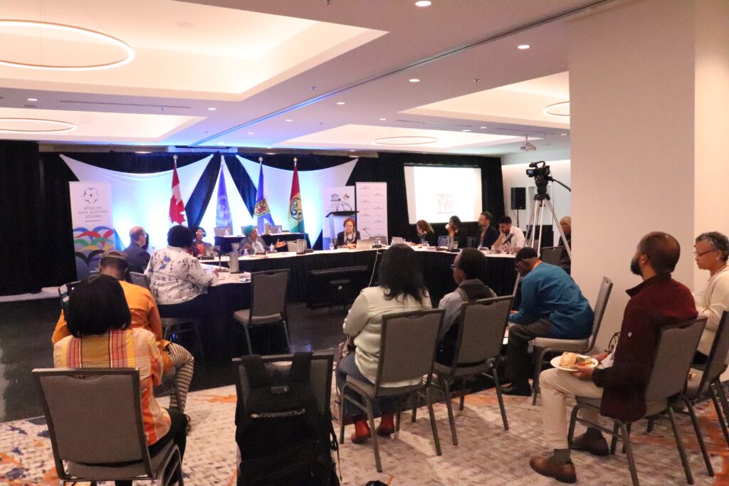 A group of UNESCO committee members seated at a round table with spectators looking on.