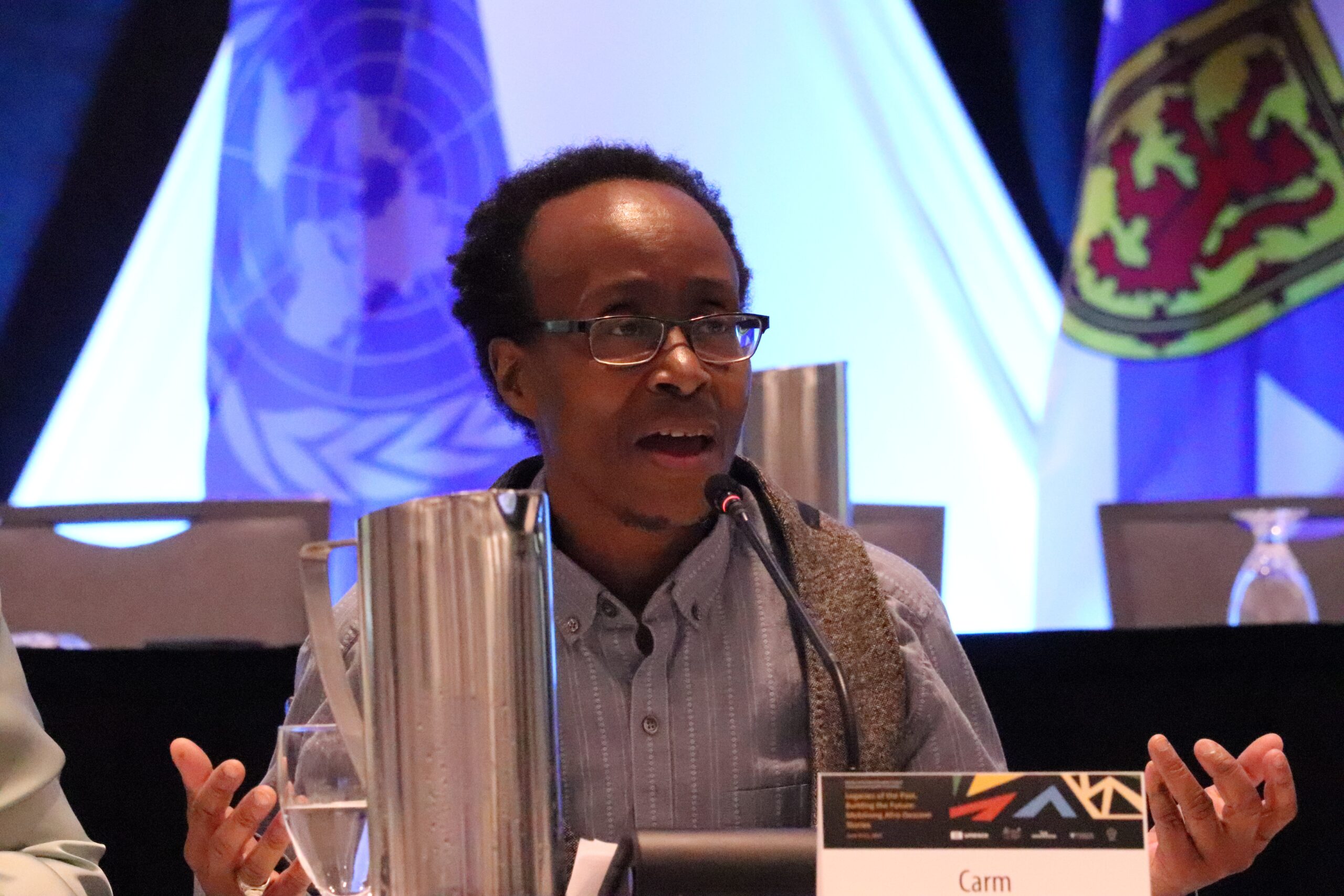 Black person in grey collar shirt and glasses speaks at a panel presentation.