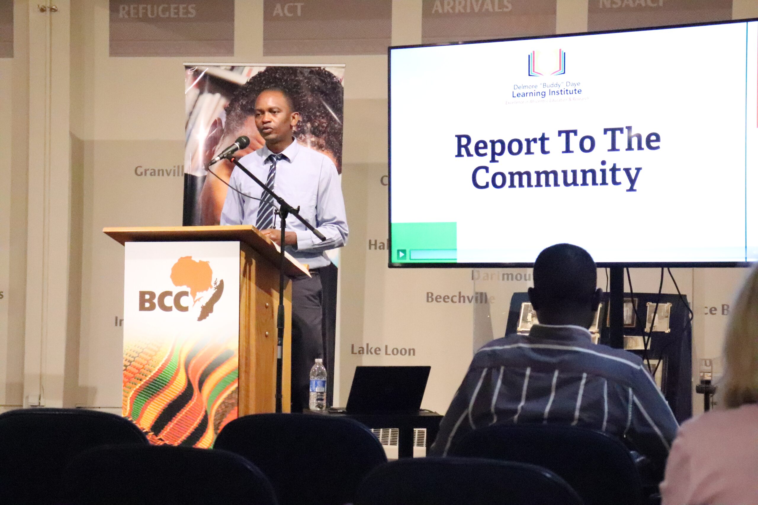 Black man in shirt and tie speaks at a podium next to a TV monitor