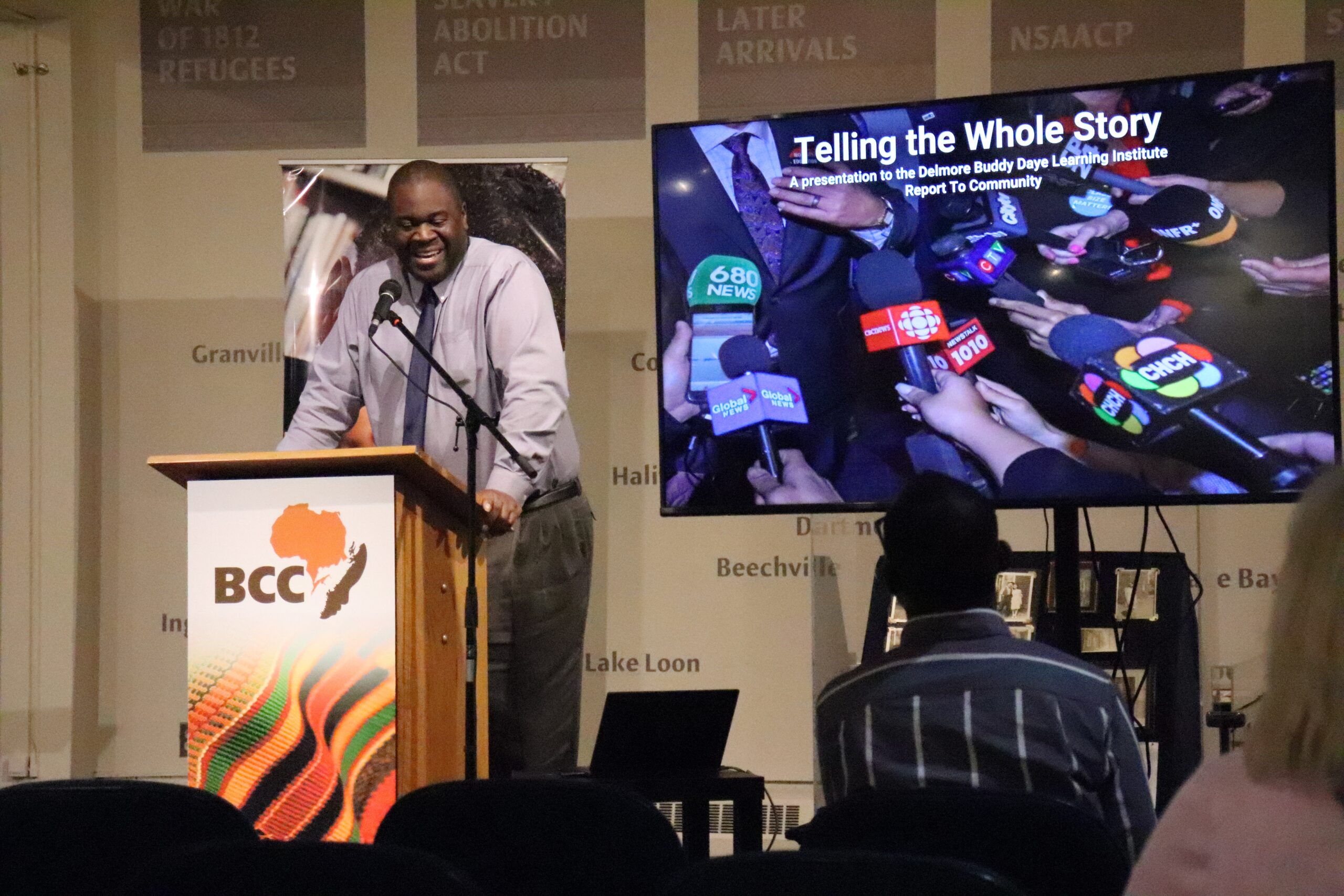 一个穿衬衫打领带的黑人在电视显示器旁的讲台上讲话