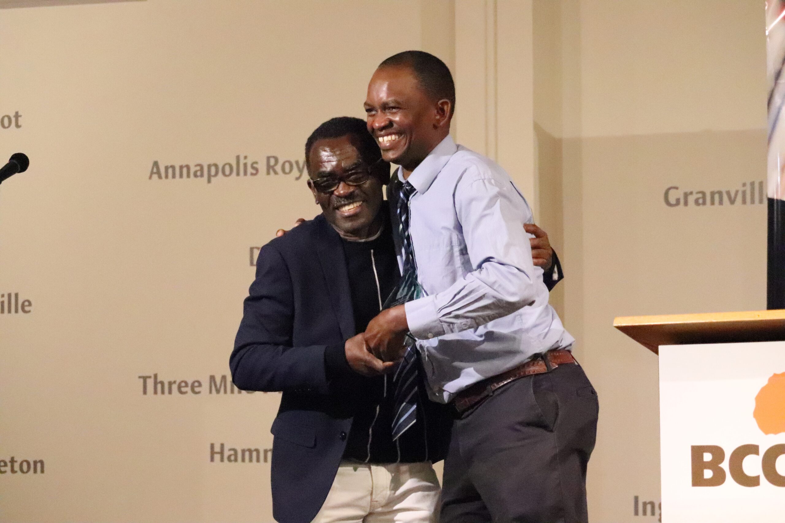 Two Black men smile to the audience while embracing each other on stage.