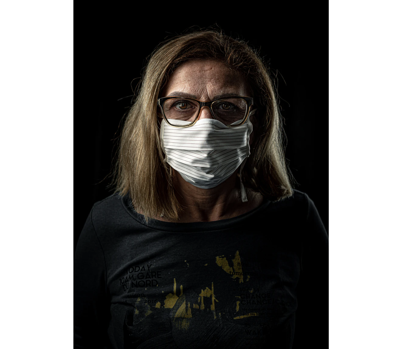 A woman in a white mask stands against a black background looking solemn over her glasses.