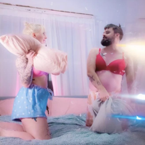 A blonde woman and a white man with a dark beard, both wearing pajama bottoms and either a red or a pink bra, have a pillow fight on a bed.