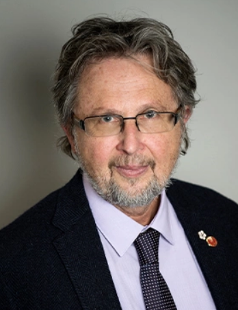 A headshot of a man with greying hair, glasses, a grey moustache and beard, wearing a dark suit, light coloured shirt, and tie.