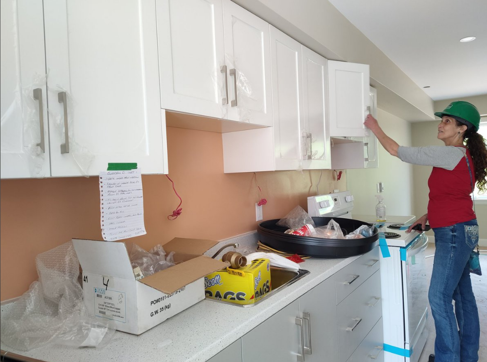 一名戴着绿色安全帽、穿着红灰t恤和牛仔裤的女商人正在一栋在建房屋内检查白色橱柜。