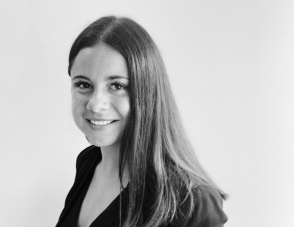 A black and white photo of a young woman with long hair, side on wearing a dark shirt and smiling at the camera.