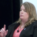 A white woman in a pink blouse raises a finger while she speaks