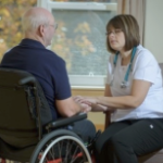 A white female nurse comforts a senior white man in a wheelchair