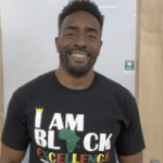 A young Black man smiles wearing a Black T-shirt which reads 