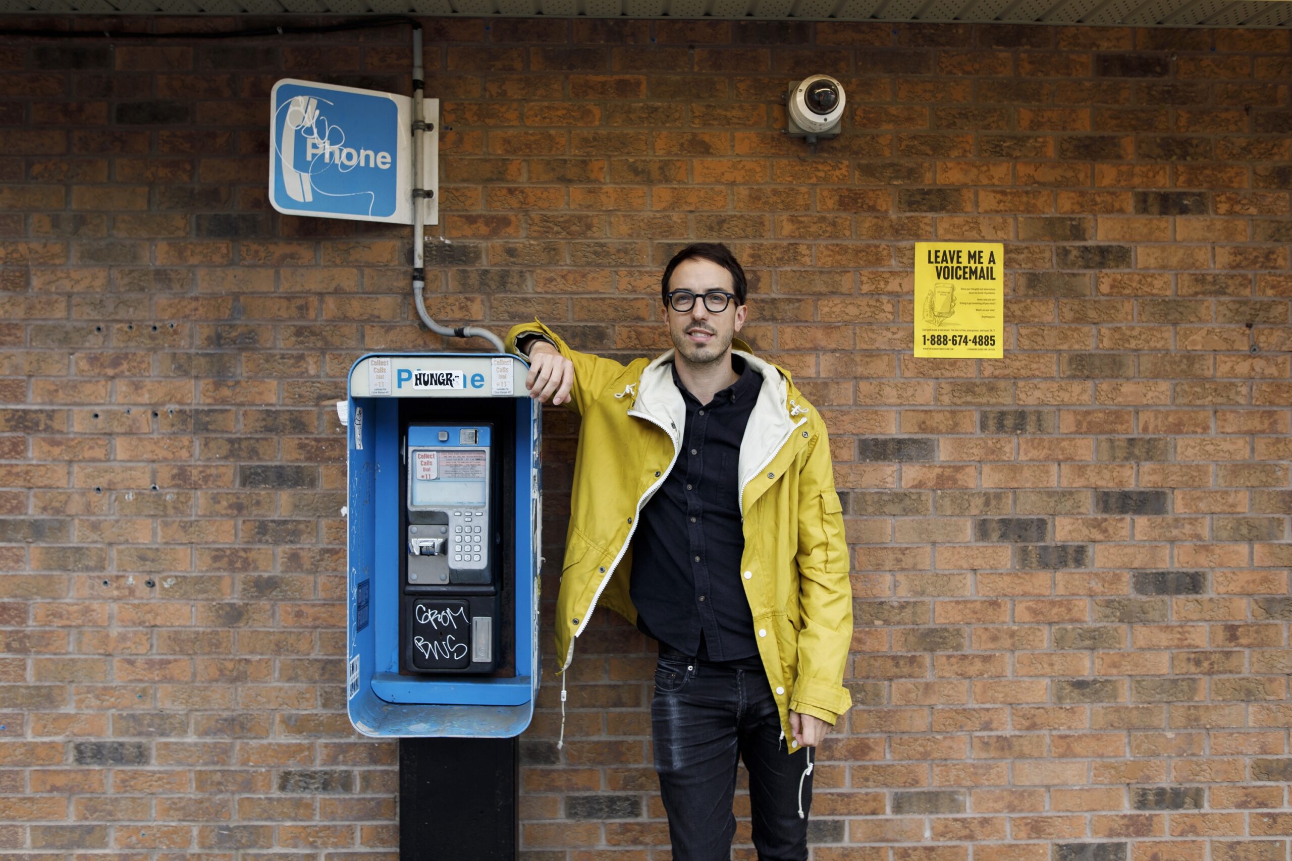 一个穿黄色夹克的白人靠在公用电话上。他身后的墙上挂着一张海报，上面写着“给我留言”。