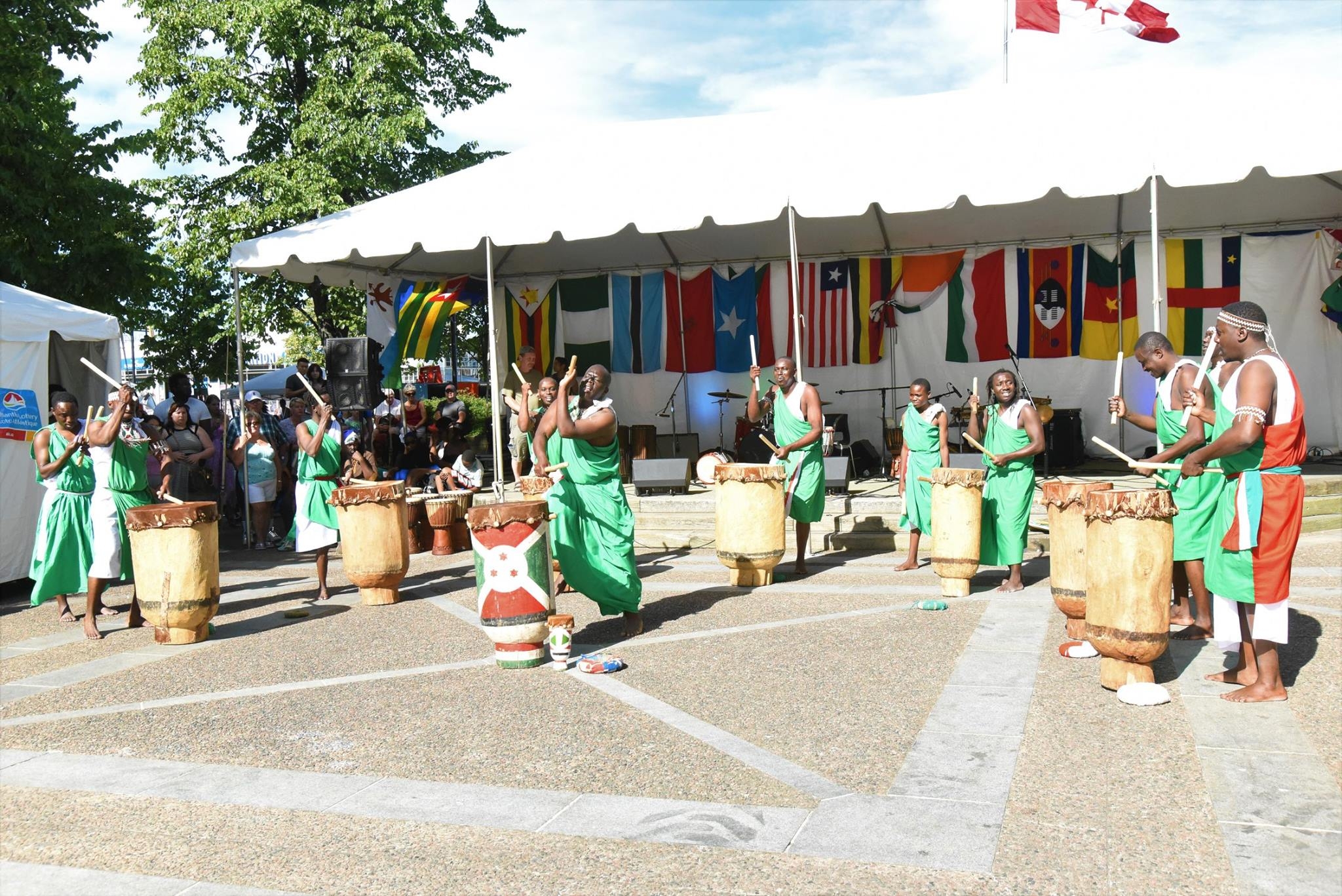 身着绿色服装的非洲鼓手在户外表演。后面的建筑有白色的遮阳篷，墙上挂着几面非洲国旗。