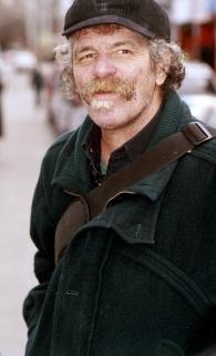 一个留着大胡子的白人站在街上。他穿着黑色夹克，戴着黑色帽子。