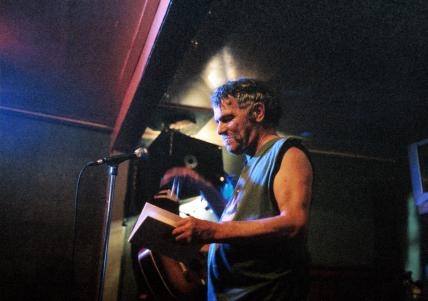 一个白头发的白人穿着无袖衬衫站在舞台上。他拿着一本书，站在麦克风前。